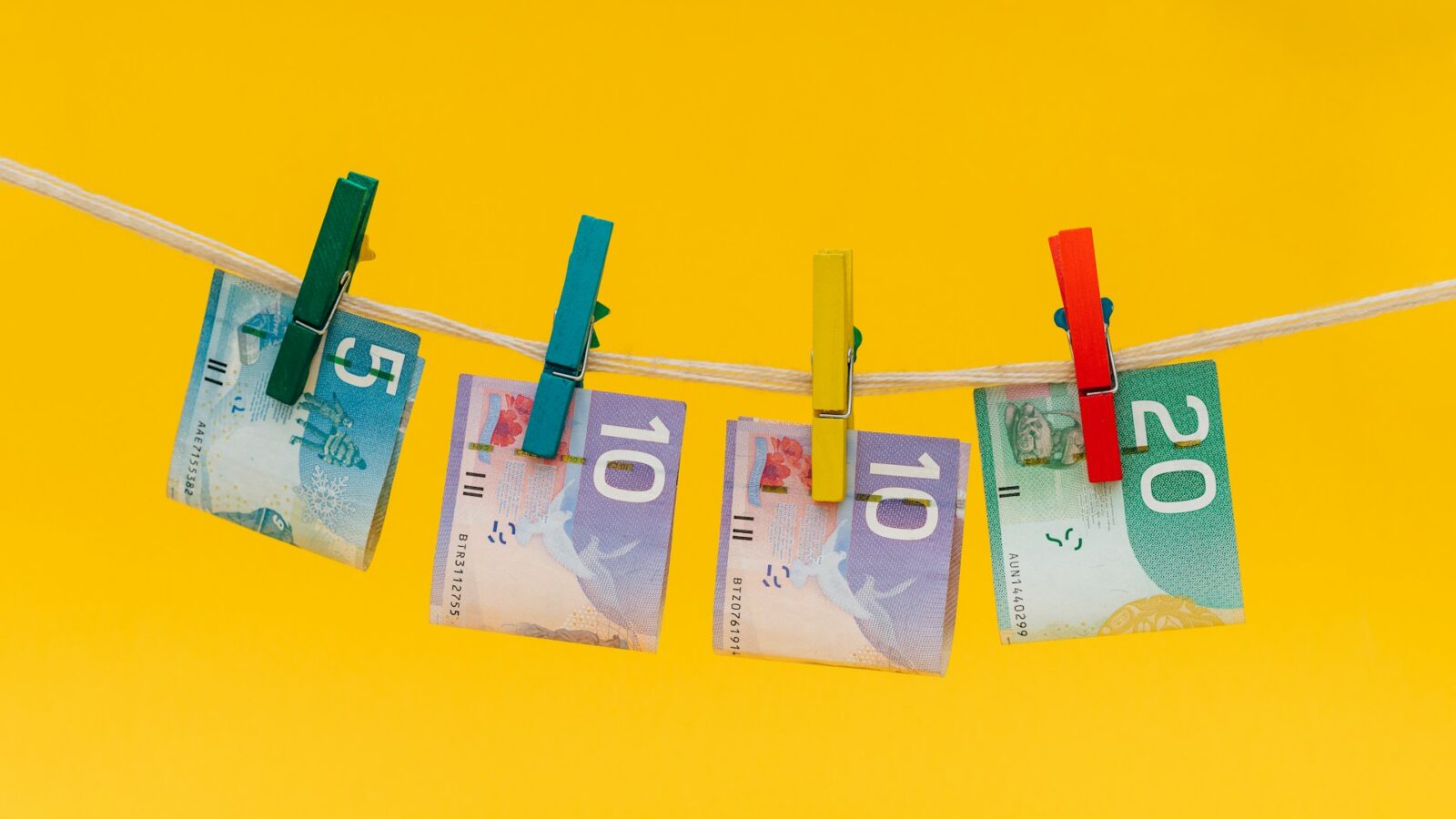 A five-dollar bill, two tens, and a twenty-dollar bill are clipped onto a laundry line with multi-colored laundry pins, representing anti-money laundering requirements.