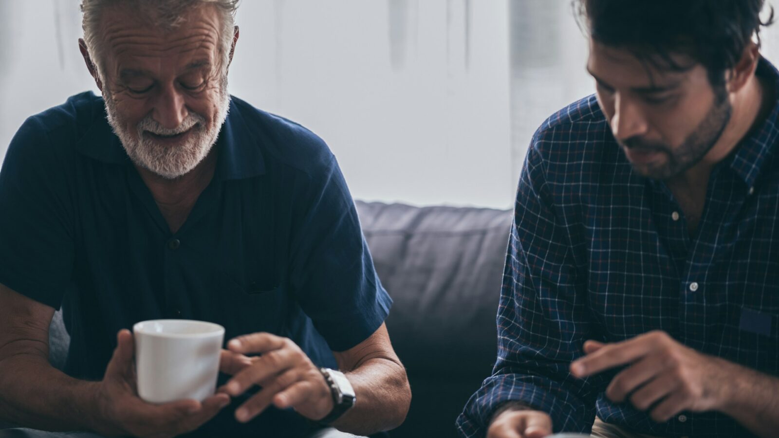A man drinks coffee with his aging father on a sofa, representing adult guardianship.