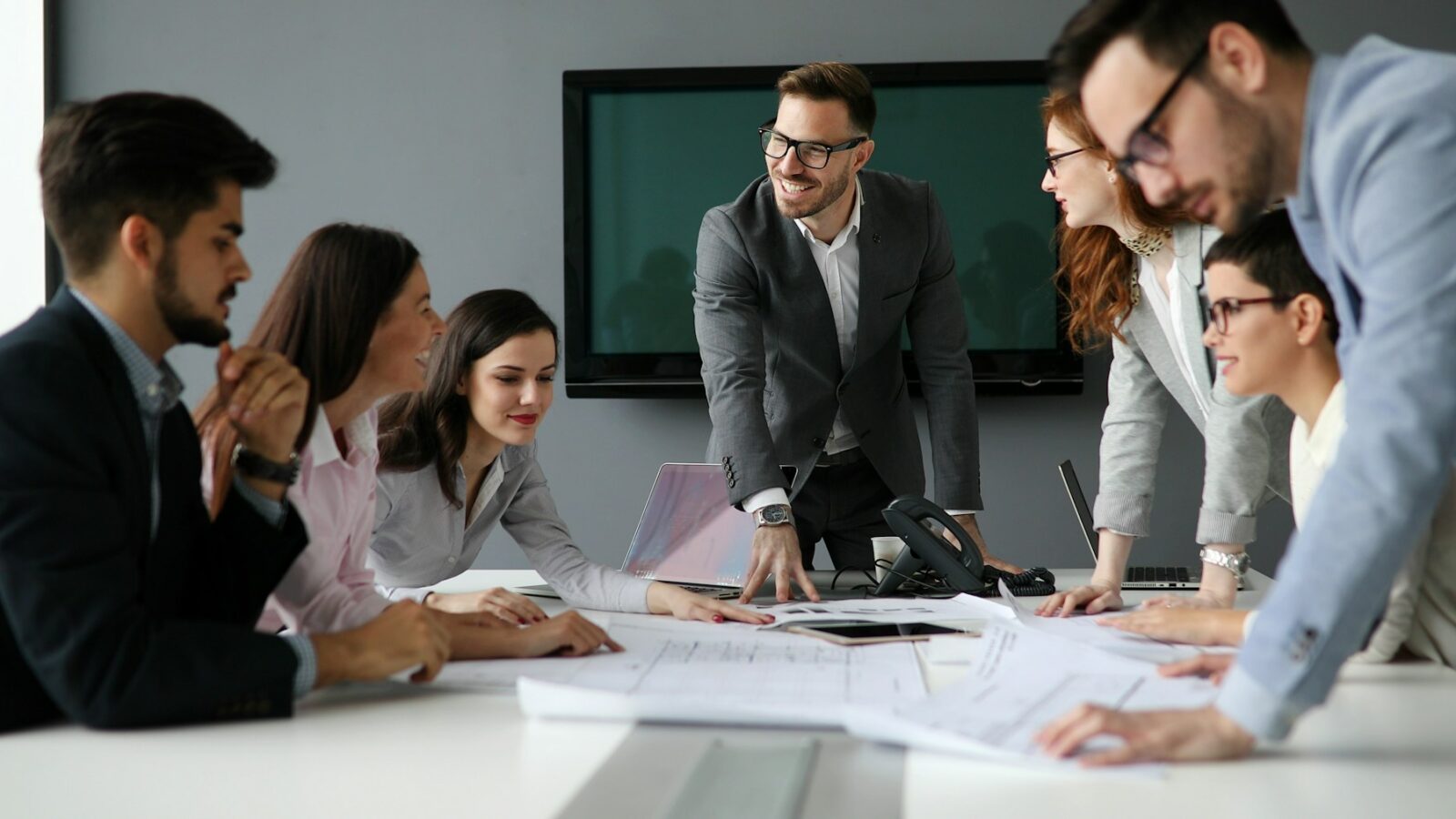 Coworkers sit and stand around a meeting table covered with paperwork, laptops, and a telephone, with a large screen television mounted on the wall behind them, representing Alberta professional corporations.