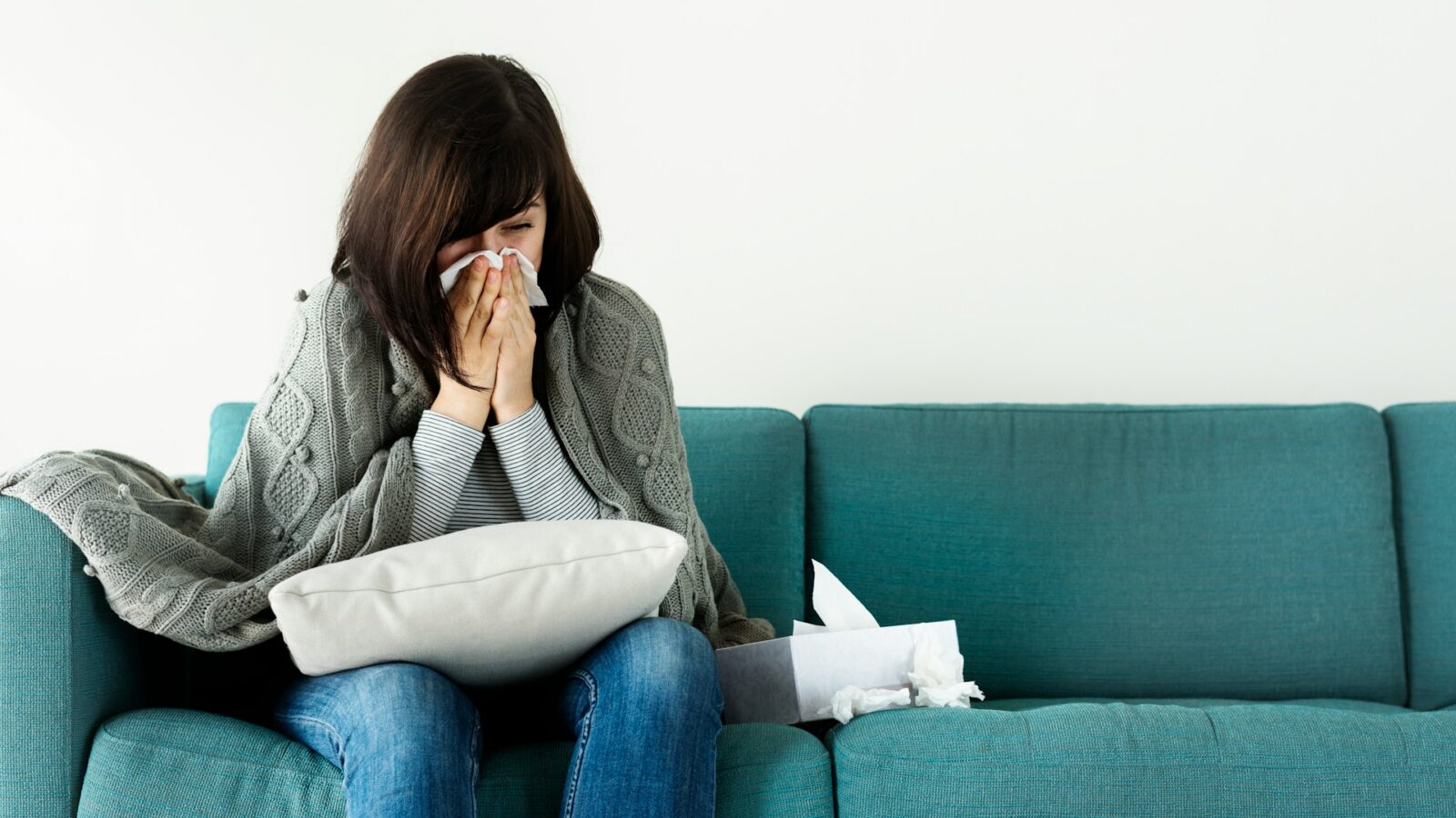 A woman sitting on a couch blowing her nose into a tissue, representing the expansion of job-protected long-term sick leave in Alberta