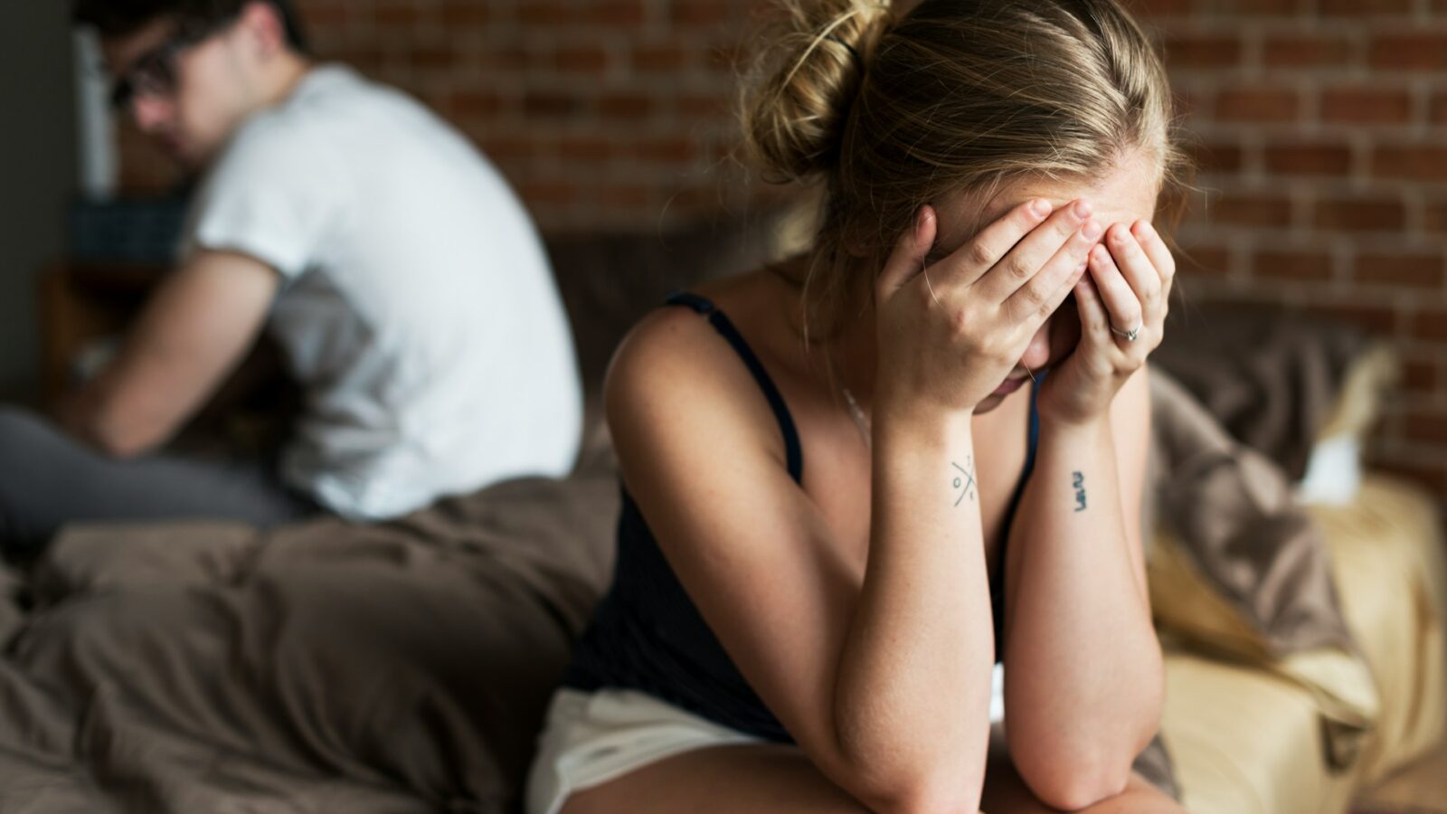 A woman sits on the edge of the bed with her face buried in her hands and her husband's blurred image sitting on the bed with his back to her, representing coercive control in Alberta family law.