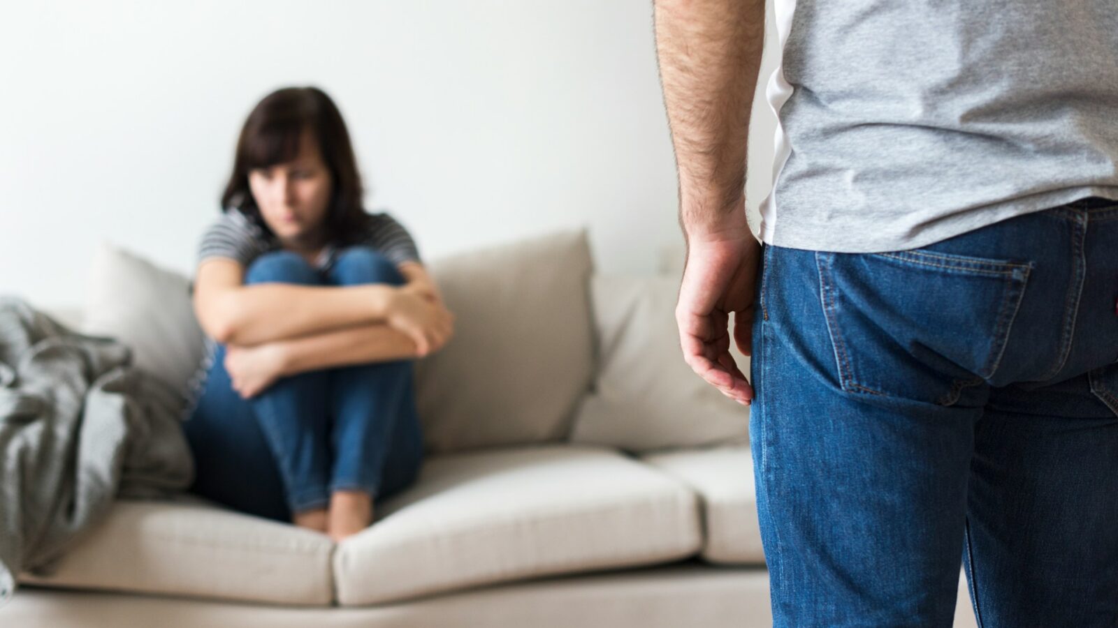 A woman sits on the sofa, curled up with her knees drawn to her chest and arms wrapped tight around her legs, unable to look at the man, visible from the elbow to his knee, standing threateningly over her, representing family law and intimate partner violence claims.