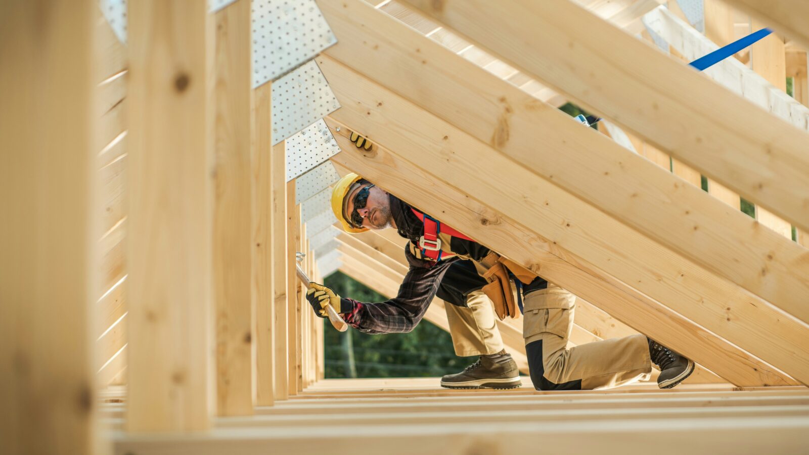 A construction worker crouches while hammering an angled roof frame, representing Alberta's construction and real estate outlook.