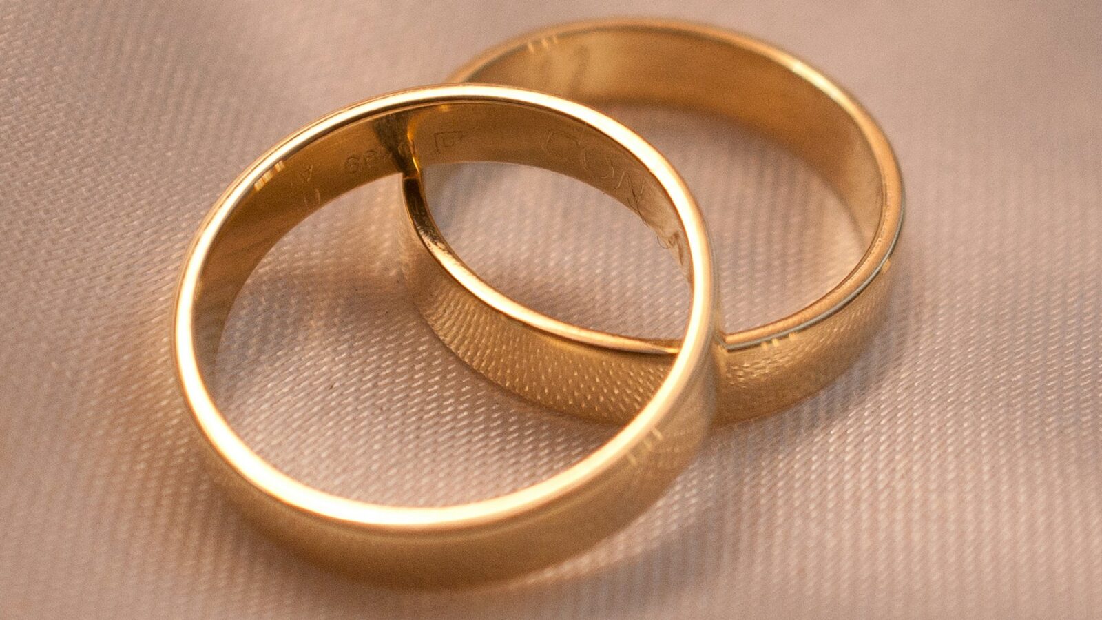 A close-up of two gold wedding bands, one leaning against the other, on beige fabric, representing marriage contracts and increased wealth after marriage.